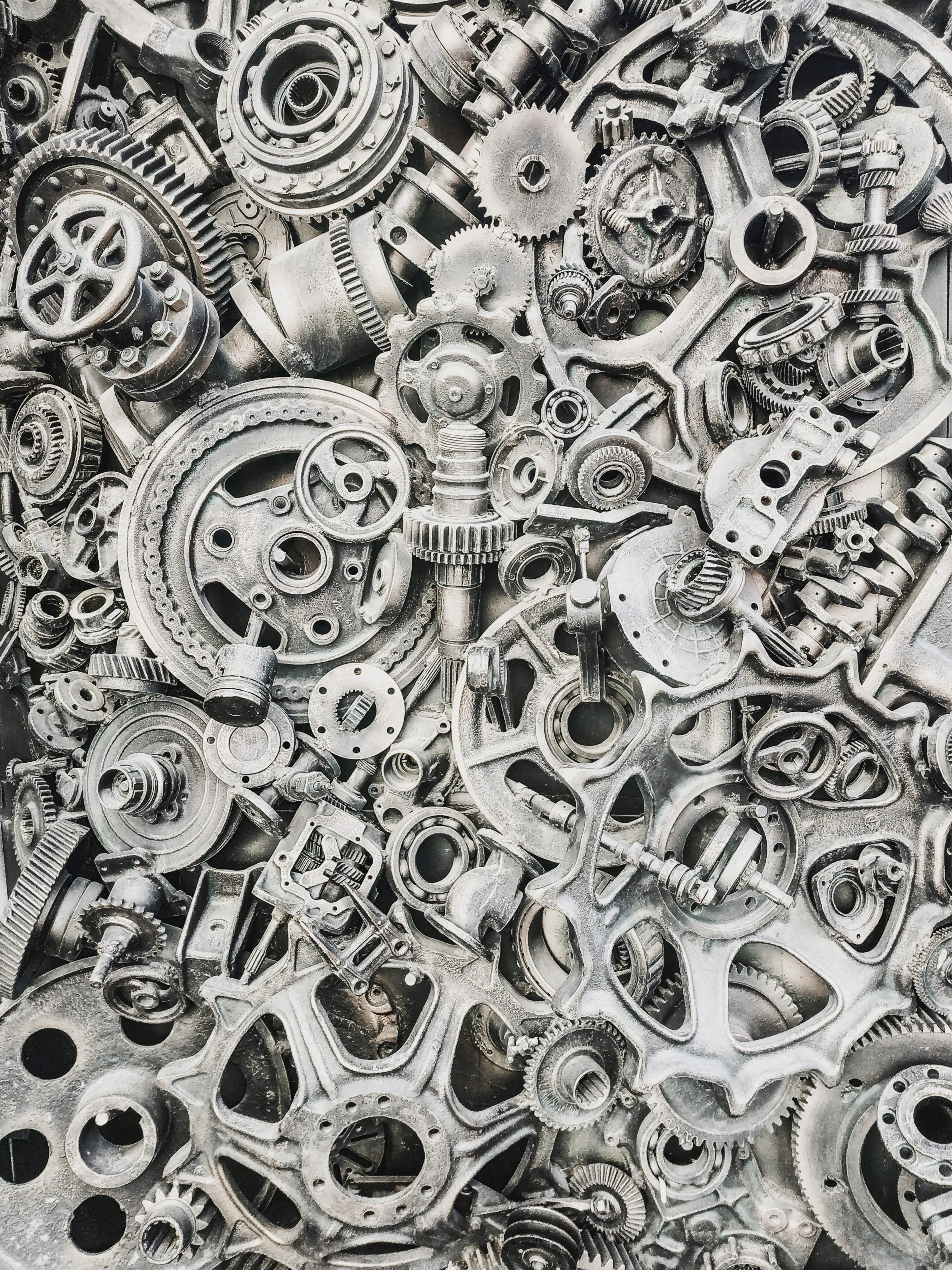 A detailed close-up of various interlocking gears and mechanical parts, intricately arranged. The components are metallic, with a slightly worn, industrial look. The composition is dense and complex, highlighting the intricate nature of mechanical engineering.